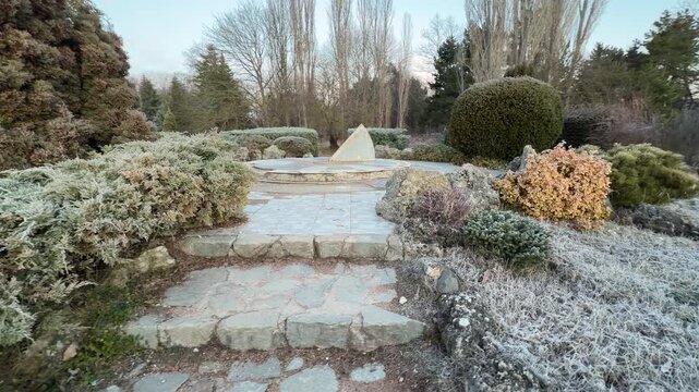 A winding stone path leads through a rugged rock garden covered in morning frost. The quiet morning light creates a peaceful and refreshing mood across the park. The air feels crisp and still.