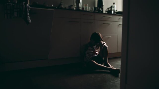 Sad child seated against kitchen cabinets with knees drawn close hiding face showing emotional suffering depression and loneliness in quiet domestic interior environment