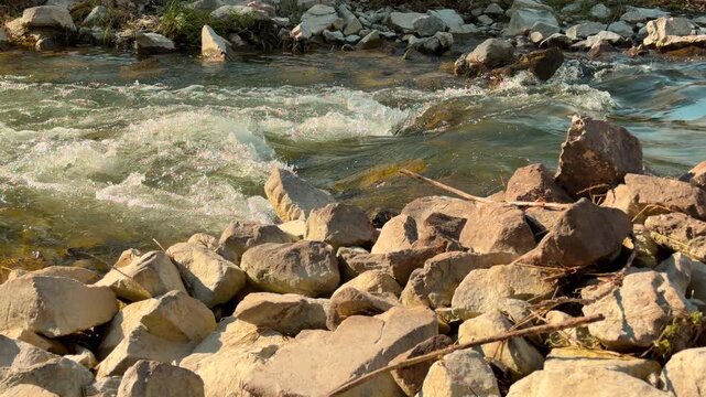 A lively mountain stream rushes over stones and creates white foam. The jagged rocks along the bank provide a sense of calm stability against the restless water. It is a refreshing and wild scene.
