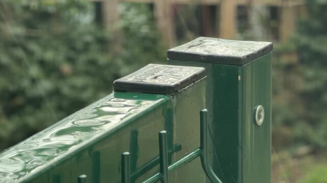 Shiny green fence posts stand still as fresh raindrops gather on their black caps. The scene feels peaceful and quiet. Soft green leaves blur in the background of this rainy day.
