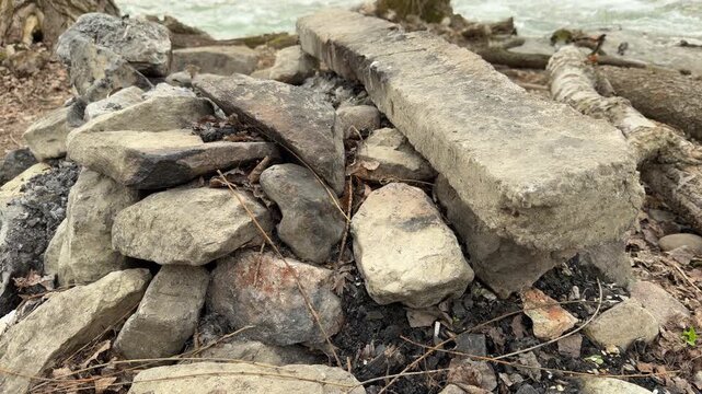 A cold, hand-built stone campfire ring sits quietly among fallen logs. The remnants of ash feel lonely against the backdrop of a fast-flowing river, evoking a sense of peaceful solitude.