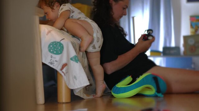 Toddler attempting to climb onto bed while holding edge but unable to reach top as caregiver sits behind handling toy showing divided attention in busy home environment
