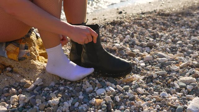 Boots socks beach person putting on black chelsea boots over white socks on a sunny pebble beach shoreline during vacation