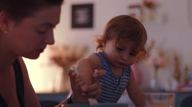 Toddler eating with spoon while sitting at table next to mother showing independence learning self feeding skills and everyday family mealtime interaction at home