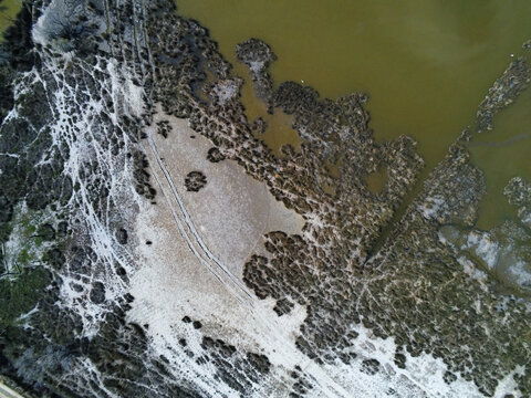 Aerial view of the rugged marshland terrain featuring salt deposits and vehicle tracks beside the murky water in Saint-Laurent-d'Aigouze, Occitanie, France.