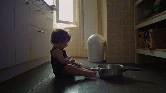 Toddler sitting on kitchen floor playing with metal pots and utensils, curious expression, exploring household objects, warm home environment, early childhood learning and play