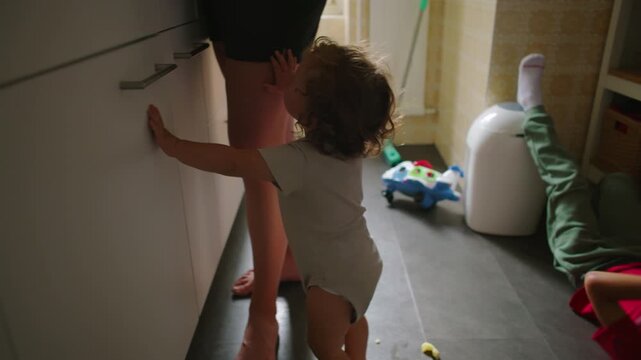 Toddler sitting on kitchen floor then standing and reaching toward mother at countertop, seeking attention while she cooks, emotional parenting moment, everyday family life and care