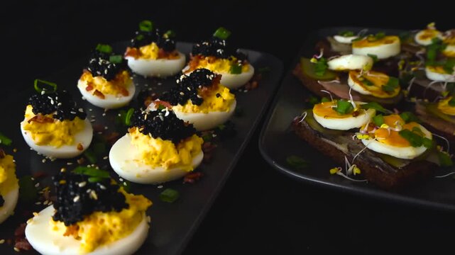 Delicious deviled eggs with black caviar and sandwiches with herring, pickles, and sprouts arranged on black plates for a celebration, party, or catered event on a dark background