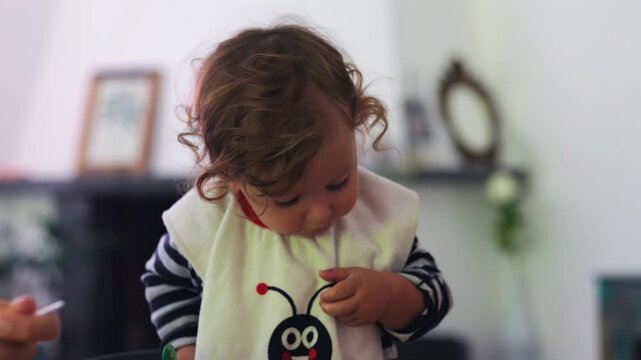 Toddler standing at table using spoon to eat from bowl while parent hand offers food nearby, active learning moment showing coordination, independence, and supportive family interaction