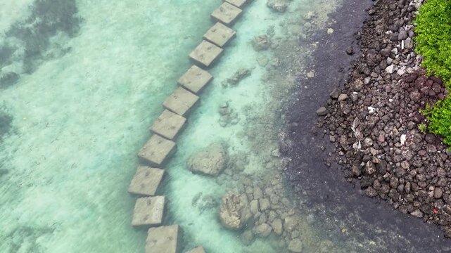 Aerial view of a unique square block barrier in crystal clear shallow ocean water by a rocky shore with green foliage.