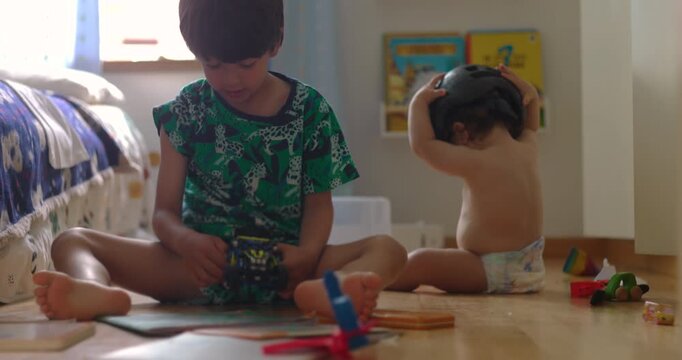 Two children sitting on floor playing independently with toys, older boy focused on vehicle while baby adjusts helmet, authentic sibling dynamic showing exploration and parallel play