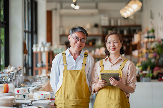 Senior Asian owner and a young female colleague smile proudly with a tablet inside their cozy homeware store full of pottery.