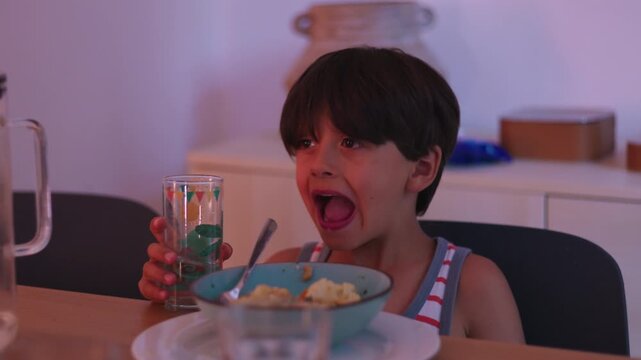 Young boy drinking water from glass while sitting at dining table during mealtime, quiet family routine showing independence, focus, and everyday childhood moment at home
