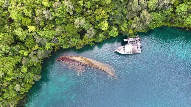 Aerial view of sunken shipwreck and diving boat in turquoise tropical water surrounded by lush jungle.