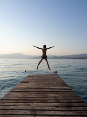 Young boy jumping from wooden pier into lake at sunset, back view of child having fun during summer vacation with mountains and calm water in background © Violeta