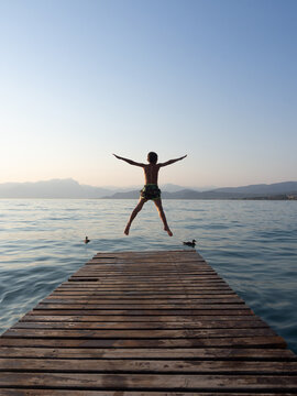Young boy jumping from wooden pier into lake at sunset, back view of child having fun during summer vacation with mountains and calm water in background