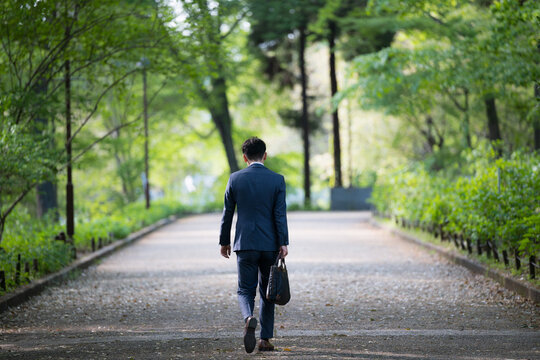 新緑の公園をビジネスバッグを持って歩く営業マンの仕事イメージ全身の広角　顔無しの後ろ姿、落ち込むネガティブストレスイメージ