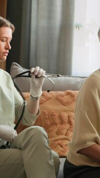 Vertical shot of young Caucasian nurse with stethoscope auscultating lungs of patient sitting on sofa during in home care