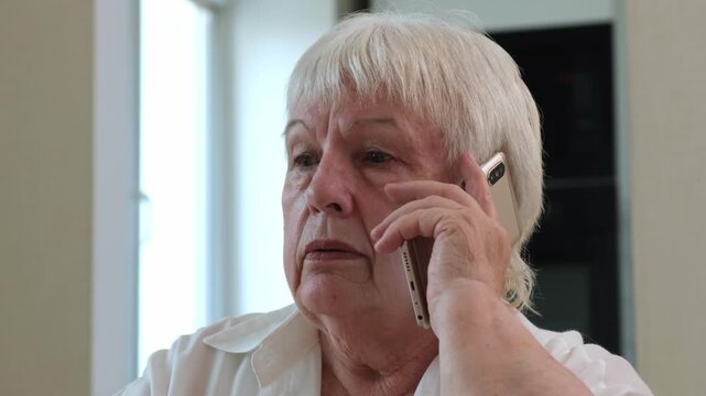 Serious senior woman talking on mobile phone at home close up. Concept of call from scammer