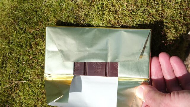 Unwrapping chocolate bar. Chocolate bar in paper-based eco-foil with a premium look, in a human hand on a green moss background. Sunlight. Ethical, natural, and organic chocolate. 