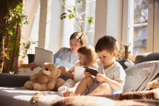 Toddler using smartphone in foreground while mother and sister interact with laptop behind. Concept of early gadget exposure, divided attention, modern parenting challenges and tech influence.