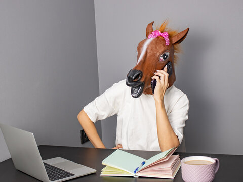 A person in a horse mask engaged in a conversation while working at a desk with a laptop, notebook, and coffee mug, showcasing a humorous take on remote work life.