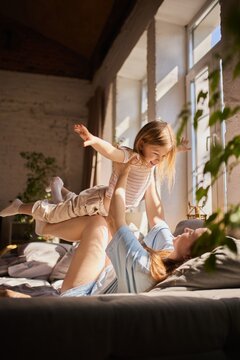 Mother playing airplane game lifting daughter on legs in sunny living room. Concept of family lifestyle promotion, parenting services, emotional wellbeing and joyful childhood moments.