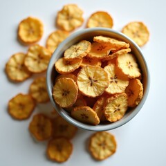 Fototapeta premium Dried banana chips are arranged in a grey bowl on a clean white surface. Slices are thin and golden brown. These healthy snacks offer a sweet taste and crunchy texture.