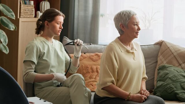 Medium long stab shot of female Caucasian medical worker listening to breath of aged woman with stethoscope during home checkup