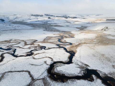 Aerial view of a meandering river winding through snow-covered fields and rolling hills in the countryside of Nasbinals, Occitanie, France.