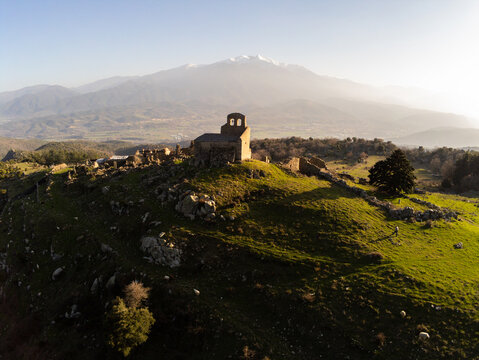 Aerial view of the ruins of the village of Comes and the church of Saint-Etienne de Comes on a grassy hill with the snow-capped Pyrenees mountains in the background Eus, Occitanie, France.