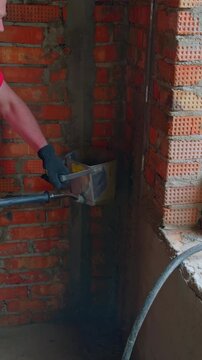A man using a hopper spray gun to apply a layer of cement mortar onto a red brick wall indoors. Construction worker applying plaster to a brick wall. Vertical video.