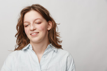 Fototapeta premium Young woman with light brown hair and white shirt looks down with a gentle smile on a plain light background. Concept of natural beauty, calmness, and casual lifestyle.
