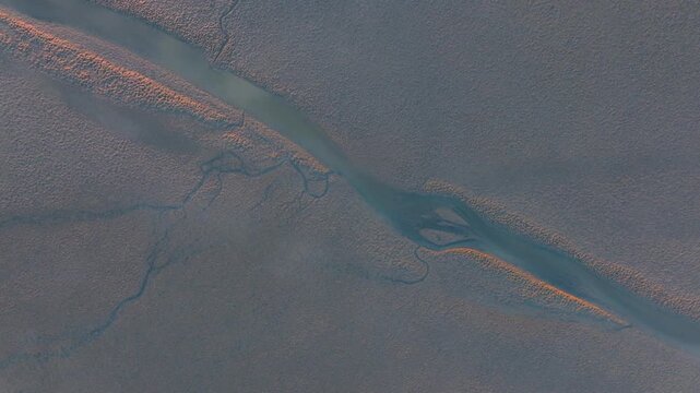 Aerial view of a tidal mudflat with winding channels at sunrise, showing the textured patterns of the exposed seabed. The low sun casts a glow on the landscape. Kawhia Harbour, Waikato, New Zealand