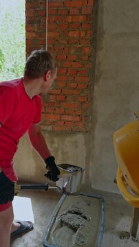 A man in a red t-shirt holding a metal spray hopper gun over a black plastic tub with wet plaster mix near a brick wall. Worker filling plaster hopper. Vertical video.