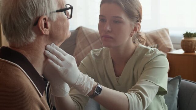 Handheld shot of Caucasian female healthcare worker examining neck of elderly man and filling out medical card during home care
