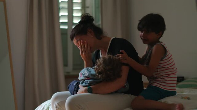 Mother sitting on bed holding toddler while covering face in emotional exhaustion as older child cries and leans on her showing overwhelming parenting stress and family pressure