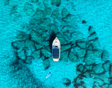 Aerial drone view of white boat anchored above rocky seabed in shallow turquoise sea, paddleboard floating nearby, calm summer scene in Mallorca, Balearic Islands, Spain. Top down view of yacht