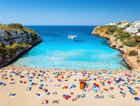 Aerial view of colorful umbrellas on crowded sandy beach, boats, hotel, rocks, swimmers in blue sea, green trees on sunny summer day. Mallorca, Balearic Islands, Spain. Tropical top drone view. Travel