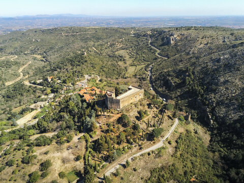 Aerial view of the medieval Castle of Castelnou perched on a rocky ridge surrounded by lush green hills and winding roads under a clear blue sky in Castelnou, Occitanie, France.