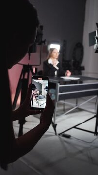 Hand holding a smartphone recording a woman sitting at a desk in a studio with lights. Backstage view of person filming video interview with smartphone. Vertical video.