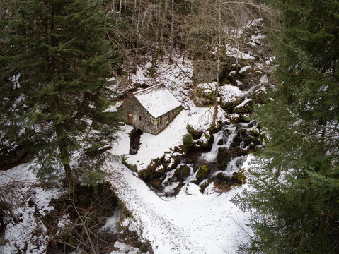 Aerial view of the Moulin de Chambeuil stone mill nestled in a snowy forest beside a cascading waterfall and stream in Laveissiere, Auvergne-Rhone-Alpes, France.