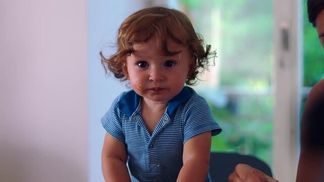 Toddler leaning forward at table with food on face during mealtime, expressive moment showing curiosity, engagement, and messy authentic childhood behavior in family setting