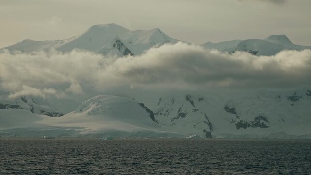 Low orographic clouds wrap dramatically around Antarctic snow-covered mountains viewed from expedition ship on dark choppy ocean