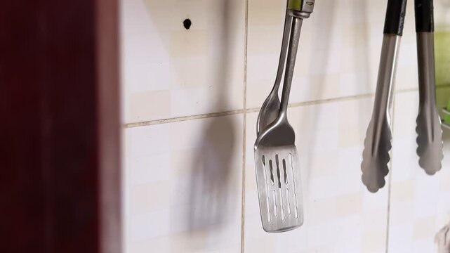 hanging metal spatula and kitchen tongs against a tiled wall in a home kitchen, with soft indoor light and subtle utensil movement.