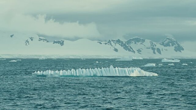 Rare spiked Antarctic iceberg with dramatic row of ice pinnacles floating in teal choppy ocean before snow-covered mountains