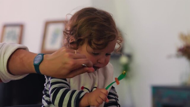 Toddler practicing self feeding with spoon while parent hand gently supports nearby, natural mealtime progression showing growing independence, coordination, and patient guidance at home
