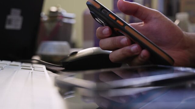 a hand holding a smartphone at an office desk beside a keyboard, mouse, and another phone, captured indoors with soft light and shallow depth of field.