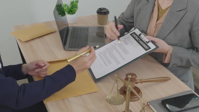 Two professional women reviewing and signing documents in an office with legal symbols, representing legal consultation, business agreement, and contract signing.