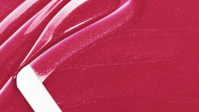 Close up of metal spatula playing with red lip gloss texture, satisfying lipgloss texture stroke. Pink cosmetic product swatch background